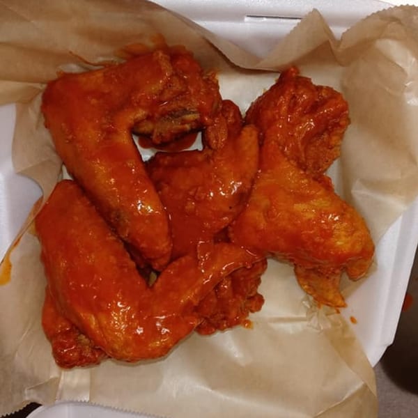 Several saucy, red chicken wings from The Blazing Chicken Shack II in a white takeout container.