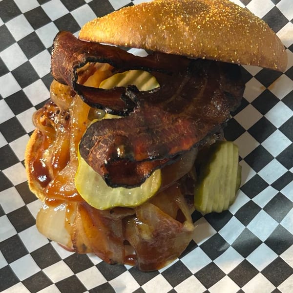 A Blazing Chicken Shack II sandwich with bacon, pickles, and caramelized onions on a checkered paper.
